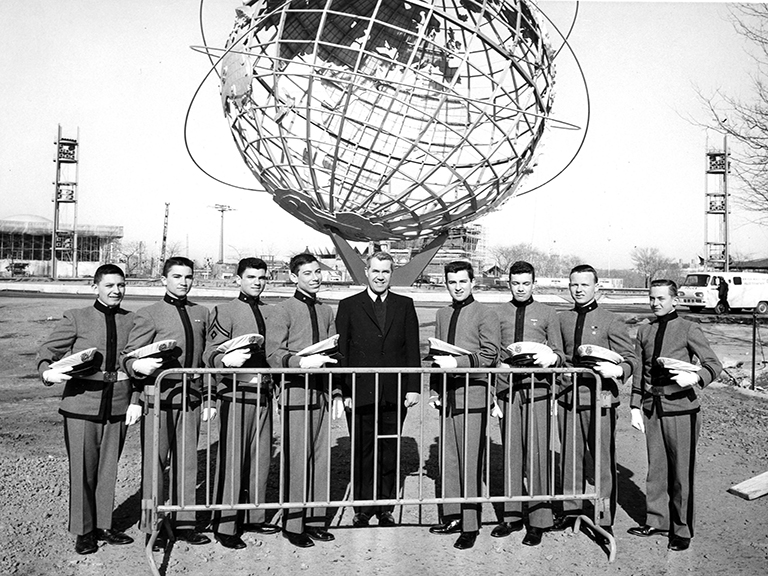Eight student cadets dressed in uniform and their assistant headmaster, a Christian Brother, line up behind a metal gate and in front of the large, stainless steel Unisphere in Flushing Meadows Park.
